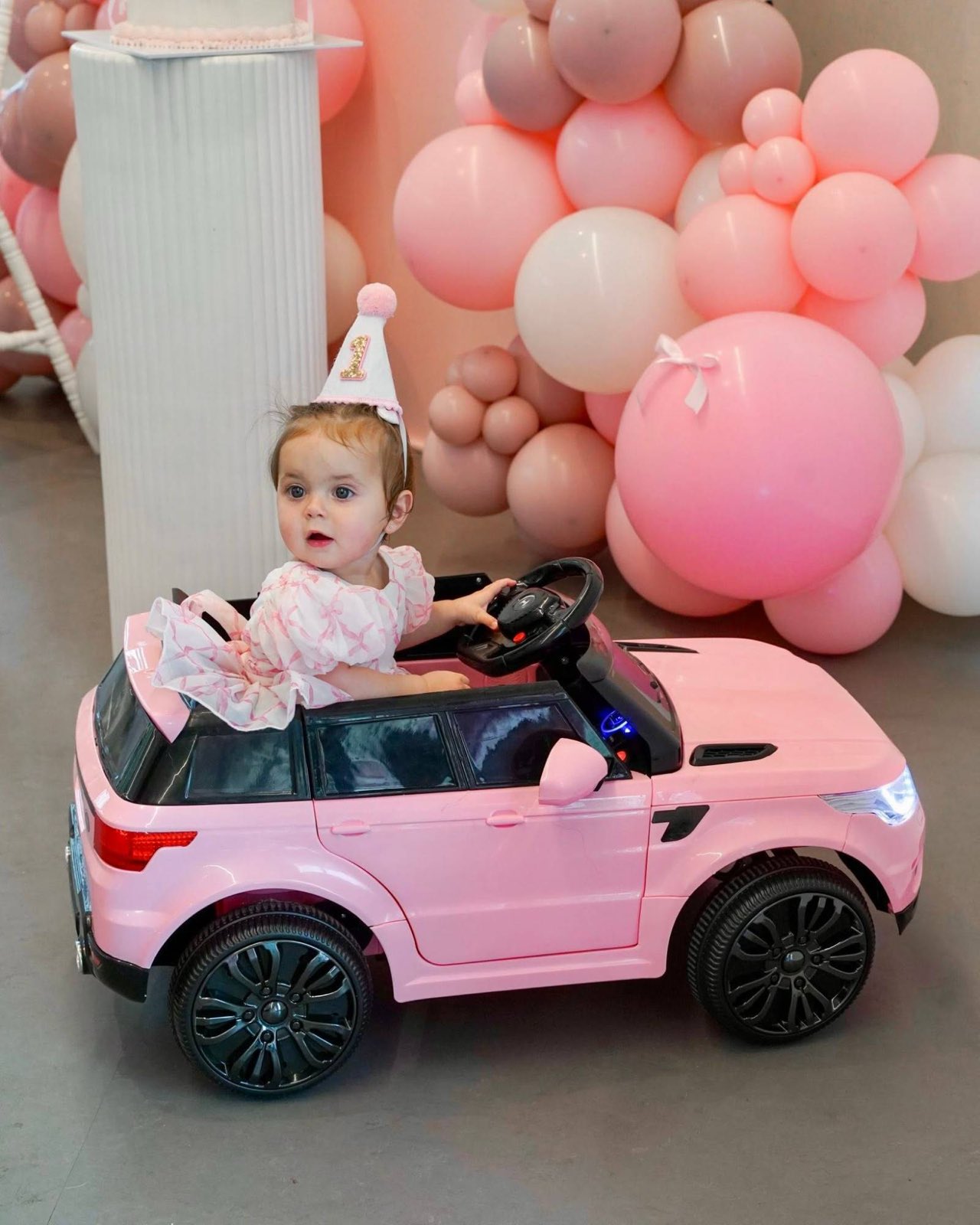A little girl in a party hat driving a pink toy car at her first birthday